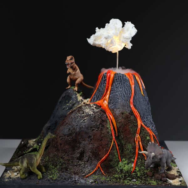 קארין עמנואל עם עוגת הר געש תלת מימד עם עשן ועננים ועליה דינוזאורים טי-רקס טריצרטופס דיפלודוקוס מפוסלים בבצק סוכר