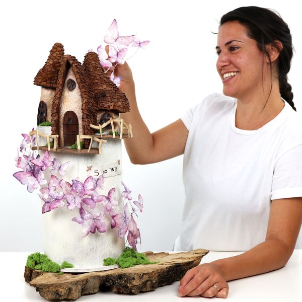 קארין עמנואל עם עוגה מעוצבת סופר גבוהה מצופה בטקסטורת שוקולד עם פסלים של טירה מפוסלים מבצק סוכר ופרפרים מנייר אורז